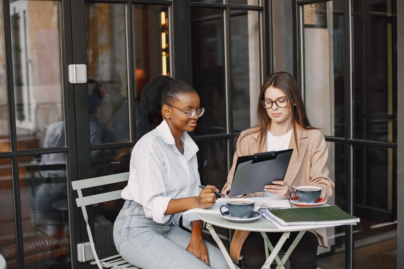 Two women talking at a cafe table with notebooks and coffee
