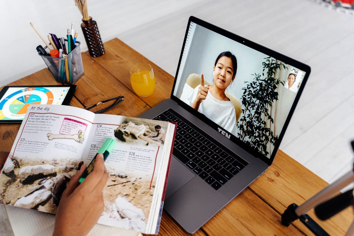 An open textbook and a laptop showing an online class on a wooden desk