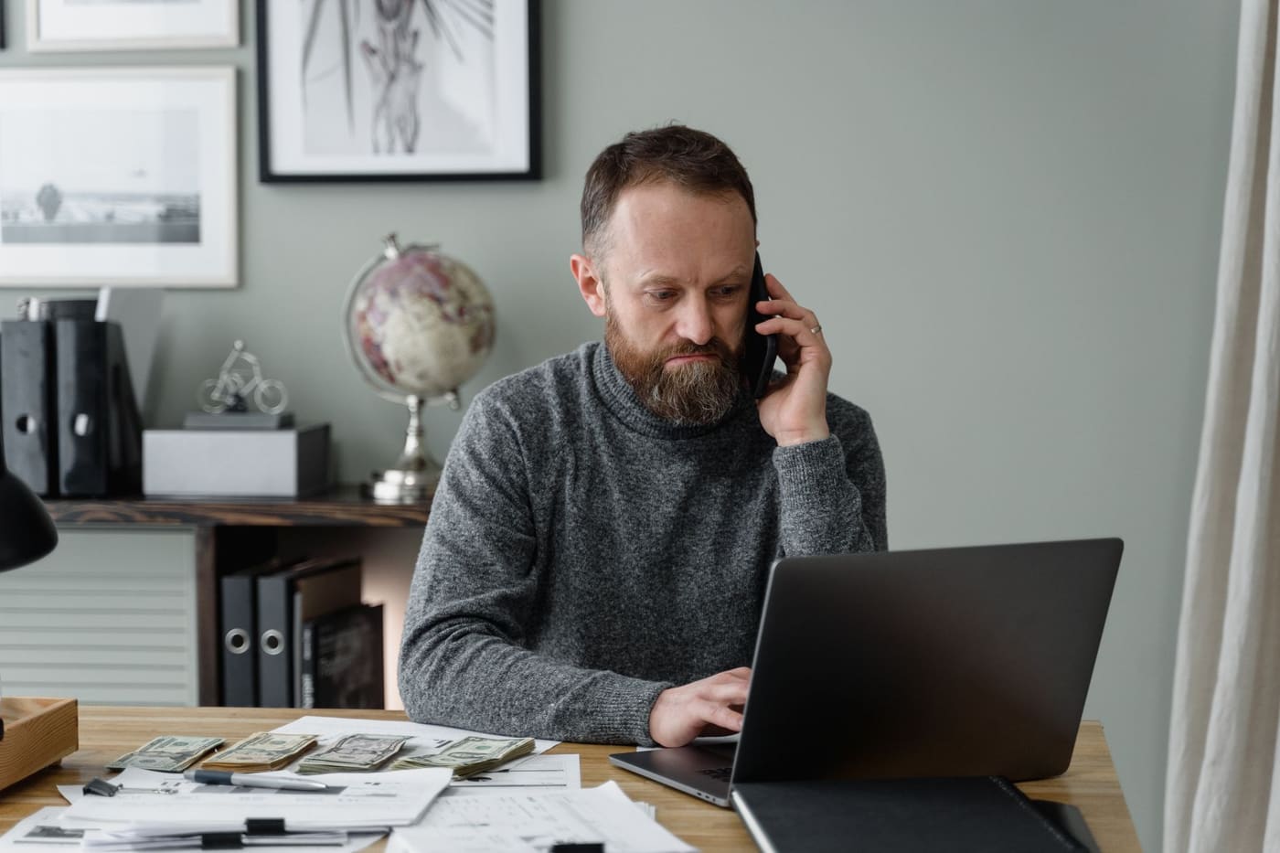 A man in a gray sweater on a phone call at a laptop desk